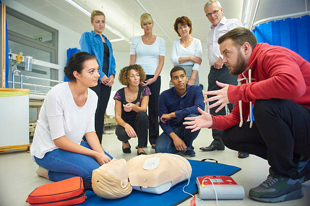 a mixed age group listen to their tutor as he shows the procedure involved to resuscitate using a defibrillator