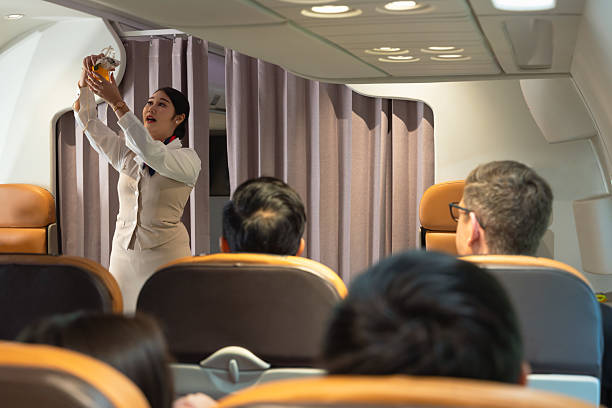 Assistente di volo/Cabin crew che sta facendo una dimostrazione di sicurezza ai passeggeri che la guardano attenti, seduti nei loro sedili di aereo, e lei sta mostrando come funziona una maschera di ossigeno.