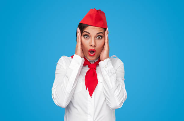 Assistente di volo stupita in uniforme elegante, con le mani vicino al viso e la bocca aperta, mentre guarda verso la fotocamera su sfondo blu.
